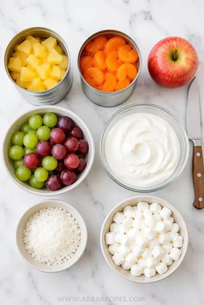 Ingredients for creamy fruit salad recipe laid flat on a white marble surface including Greek yogurt, canned pineapple, mandarin oranges, grapes, apple, coconut, and marshmallows