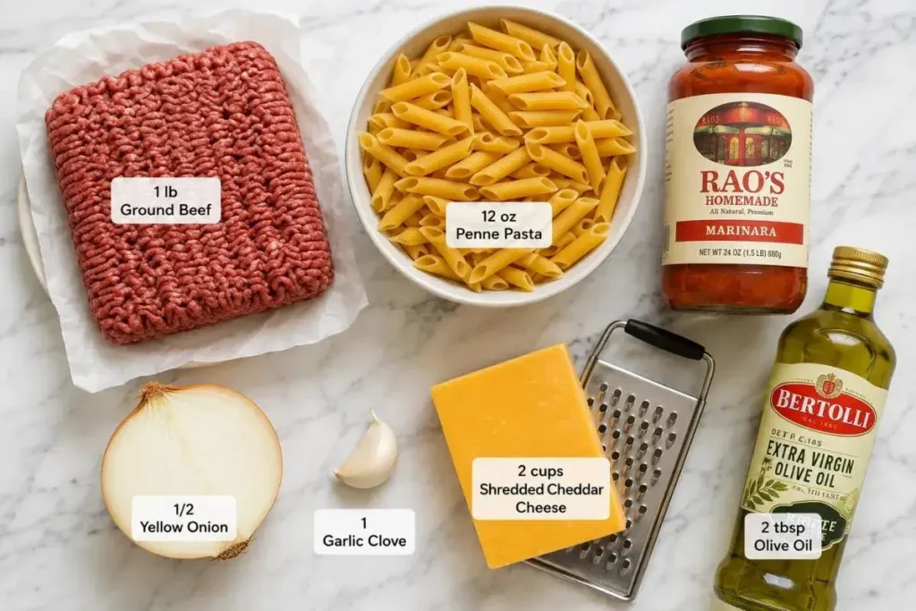 All ingredients for ground beef casserole laid out on marble: penne, ground beef, onion, garlic, marinara, and cheddar cheese